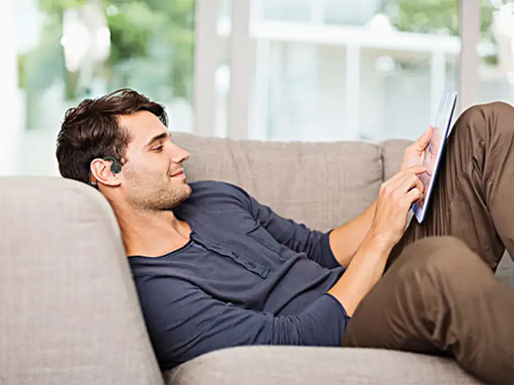 Homem relaxado usando aparelho auditivo com tablet em ambiente confortável, destacando inovação em tecnologias de assistência auditiva, saúde auditiva, bem-estar e acessibilidade.