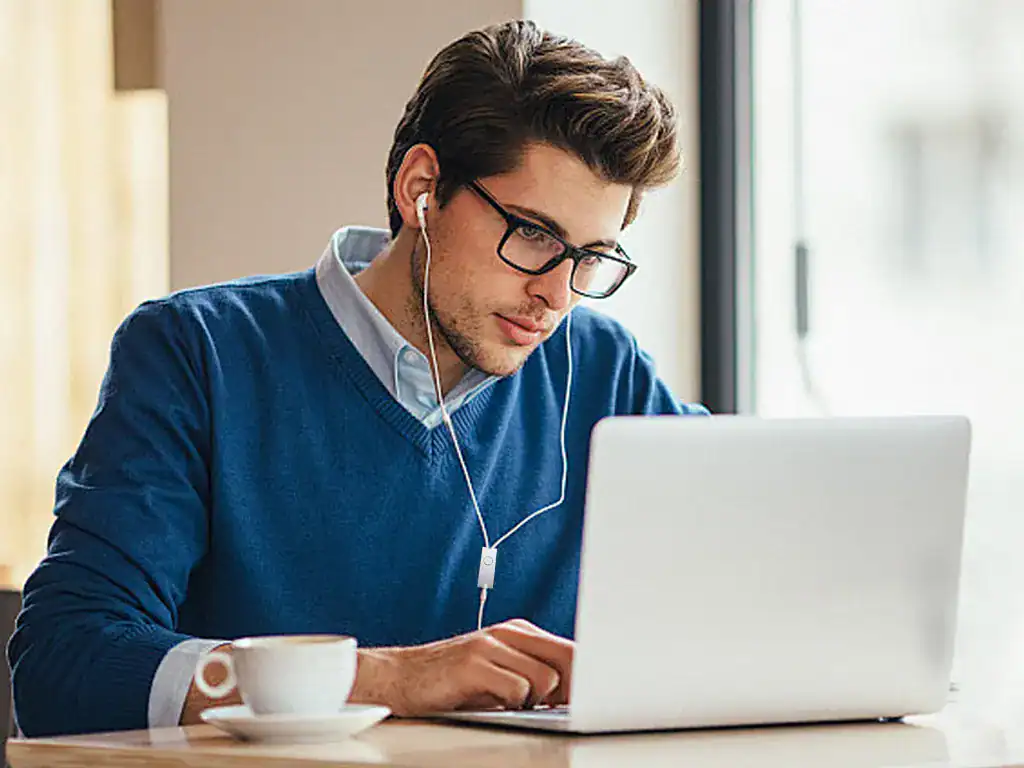 Sofá de homem jovem usando óculos e fone de ouvido em ambiente de trabalho, usando roupas casuais, conectado ao notebook, com foco na tecnologia e estilo de vida moderno.