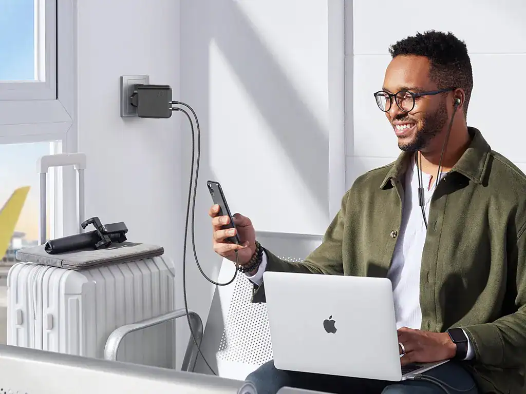 Carregador portátil conectado na parede, carregando smartphone, enquanto o homem trabalha com laptop e ouve música, na estação de trabalho moderna e tecnológica.