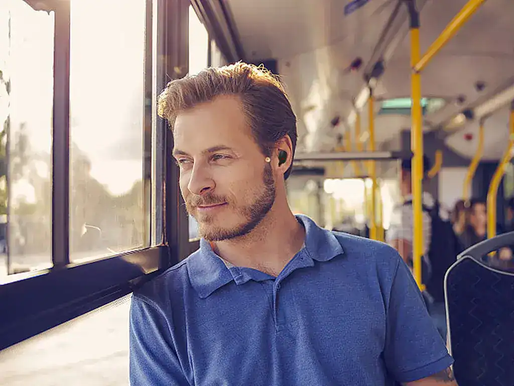 Homem usando fone de ouvido e jaqueta azul, sentado em ônibus público durante o dia, aproveitando um momento de tranquilidade e conforto ao usar um produto H'MASTON.
