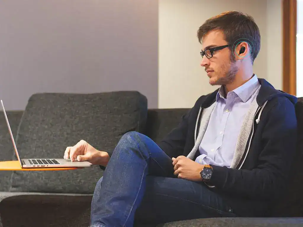 Homem jovem usando laptop com fones de ouvido em sala de estar moderna, vestindo óculos, camisa social, jeans e jaqueta, representando tecnologia, trabalho remoto, ou estudo em ambiente confortável e contemporâneo.