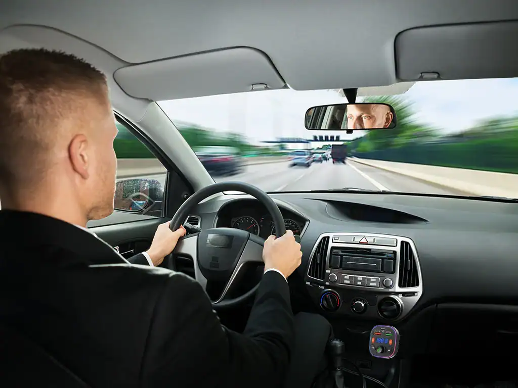 Homem dirigindo um carro na estrada, foco na direção e no espelho retrovisor, com cenário de estrada e céu ao fundo.