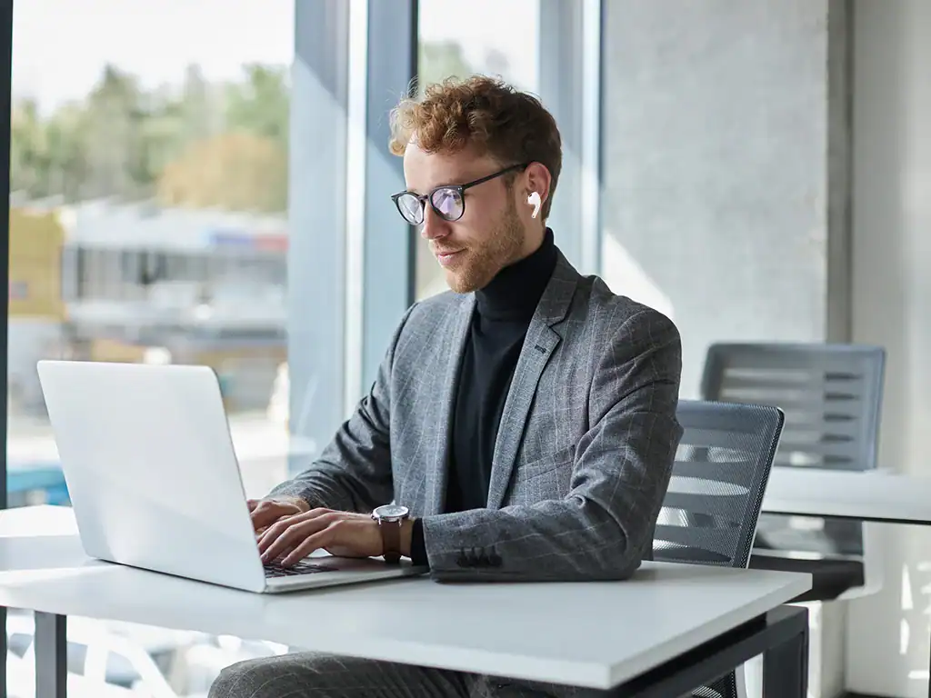 Homem de negócios com óculos, usando blazer cinza e terno preto, trabalhando em laptop em escritório com grande janela de vidro, luz natural e ambiente profissional.
